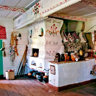 A rustic white Ukrainian wood stove fireplace with hand-painted floral and folk-art motifs, set inside a decorated room with patterned pillows, ceramic pots, and traditional wall hangings. A visual introduction to February Slavic Sunday Workshop - The Pich: Slavic Hearth Magic