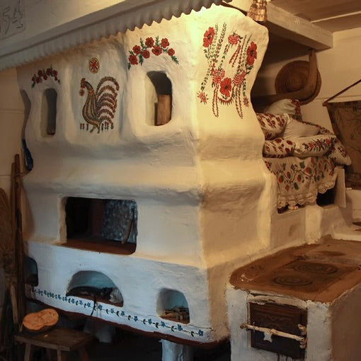 A rustic white Ukrainian wood-burning stove or fireplace with hand-painted red, green, and brown floral and rooster motifs on the front, shown in a dimly lit indoor setting.  A visual introduction to February Slavic Sunday Workshop - The Pich: Slavic Hearth Magic