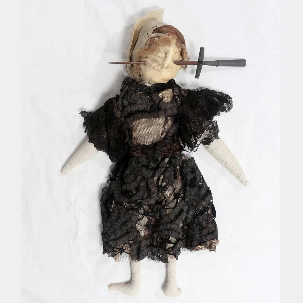 A rag doll with a beige head, black lace dress, and white fabric limbs, lying on a white background with a knife inserted into its head. Visual introduction to January Full Moon Workshop - Mastering Magical Poppets