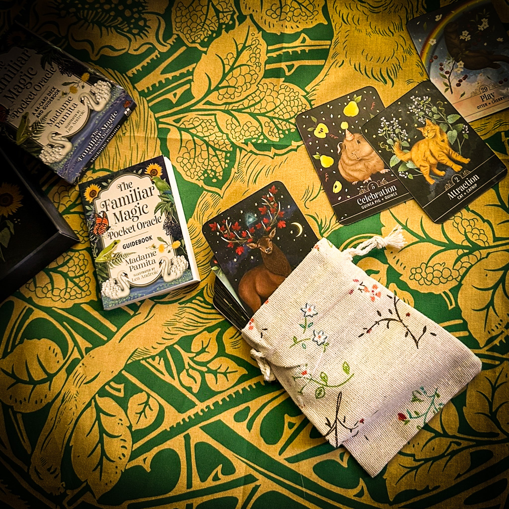A styled flat-lay of a card deck set on a green and gold patterned tablecloth. Visible items include a boxed deck, a small book titled 'The Familiar Magic Pocket Oracle', several illustrated oracle cards, and a cream fabric drawstring pouch with floral embroidery.